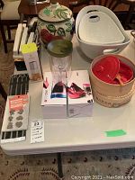 Full lot view on table showing Brie baker, ceramic jar with fruit design, white ceramic bowls, cutlery set, oil mister, Vino 2Go glasses in boxes, magnet set.