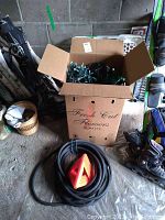 Box of Christmas lights partially unpacked with garden hose and orange nozzle coiled beside it, showing storage condition and set contents