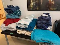 Photo of various folded bed linens including comforters, sheets and blankets piled on a table in different colors and patterns.