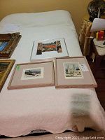Three framed lithograph prints displayed on bed: 2 smaller prints in wooden frames, 1 larger print in metal frame with matting. Printed artwork depicts human figures and abstract elements.