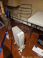 Shows two stainless steel racks and one white Westpoint oil-filled space heater on castors placed on wood floor near wall.