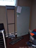 Disassembled metal frame components and rectangular glass tabletop leaning against wall, bolts and hardware on floor in plastic bags