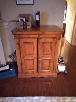 Full front view of wood TV cabinet showing two paneled doors, decorative top molding, and wooden knob handles. Cabinet stands on hardwood flooring with light wear marks on surface.
