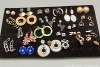 Full overview of the assorted costume earrings laid out on a black surface showing various colors, styles and materials with some pieces having missing stones.