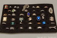 Full view of assortment of 26 costume rings on black felt display board, varying in color, style, and size, showing some missing stones and wear