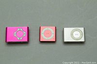 Front view of three digital music players: two Apple iPod Shuffles (pink and silver) and one Sylvania MP3 player (white), showing control pads and front colors.