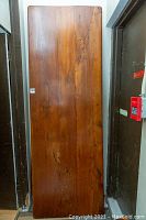 Full view of the large solid wood table top standing upright in a hallway, showing its rectangular shape and natural wood grain pattern with some knots visible.