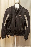 Front view of black and white Vega Technical Gear biker jacket hanging on a hanger, showing zippered front and snap closure at collar.