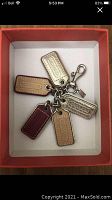 Photo showing Coach keychain inside an orange box featuring five colorful leather tags in beige, burgundy, tan, and metallic gold with Coach embossing.