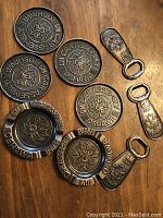 Photo showing four round metal coasters, three metal bottle openers, and two round metal ashtrays all embossed with GM Car Assembly Plant Oshawa branding featuring a maple leaf design.