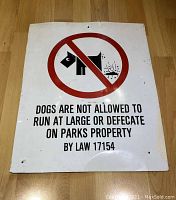 Full front view of rectangular metal sign with red circle and diagonal line symbol crossing out dog defecating, text below prohibiting dogs running at large or defecating on park property.