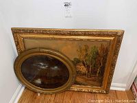 Two framed paintings leaning against wall: one large rectangular with scenic windmill, one smaller oval with ships at bay in night scene.