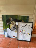 Two artworks placed together on floor: large dog painting with scratches and smaller framed abstract floral painting.