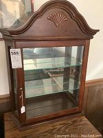 Front view of cherry wood curio cabinet showing decorative carved top, glass door with shelves inside, and brass knob.