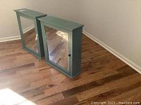 Two green wooden cabinets with glass doors placed on hardwood floor, doors closed.