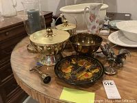 Photo showing brass lidded containers, metal rooster figurines, floral painted plate with tongs, corkscrew, candle holder, ceramic pitcher and white bowls on round table.