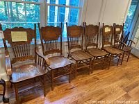 Six vintage wooden dining chairs lined up, showing four armless chairs and two armchairs with spindle back design and carved top rail, placed on hardwood floor in front of windows.