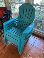 Four stackable teal green plastic Adirondack chairs photographed stacked inside a sunroom with a tiled floor near large windows showing natural light.