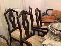 Set of six wooden dining chairs shown in corner, two armchairs and four side chairs with beige upholstery and carved backrests.