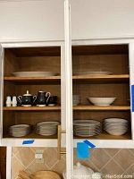 Two wooden kitchen cabinets open showing the china set inside, with plates, bowls, teapot, creamer, and sugar bowl visible on shelves.