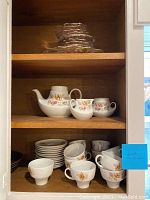 Porcelain coffee set displayed showing twelve cups, eleven saucers, coffee pot, creamer, and sugar bowl with floral gold and peach pattern, all on wood shelf.