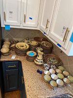 Photo showing full lot with various ceramic plates stacked, cups, saucers, covered dish, serving pieces arranged on kitchen counter.