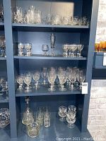 Four shelves filled with multiple sizes of stemmed glasses, decanter with stopper, glass bowl, and ice bucket in a dark blue shelving unit.