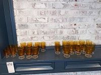 Wide view showing the full set of 27 amber glassware pieces arranged on a blue shelf against a white brick wall. Includes both footed goblets and smaller glasses in total.