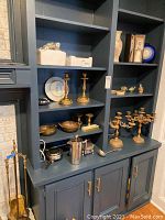 Wide view of built-in blue shelving unit with brass candlesticks, fireplace tools, candelabras, ceramic bird figurine packaging, decorative plates, and vases on shelves