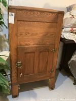 Full view of the oak ice box showing the front door with two metal latches, paneled oak wood construction, and casters on legs.