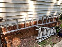 Wide shot showing the 16-foot aluminum extension ladder resting horizontally along a house exterior wall and the folded aluminum folding ladder on the ground.