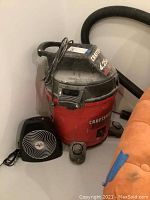 Photo showing a red and black Craftsman wet/dry vacuum cleaner with attached hose. A small black Coronado table top fan is positioned in front of the vacuum tank. The vacuum has visible dust and signs of use.