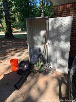 Photo of large beige Suncast storage cabinet with open doors showing interior, green and black Green Works pressure washer, black blower/vacuum parts and accessories, orange bucket and rake