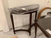 Front view of cherry demilune table wrapped in plastic showing three tapered legs and curved stretcher base.