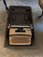 Smith Corona electric typewriter in beige color within an open brown plastic carrying case. Coiled power cord visible inside typewriter.