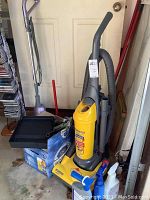 Yellow Eureka MaxIMA upright vacuum together with cleaning supplies and boxes containing light bulbs and accessories in garage area.