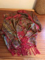 Folded 100% silk scarf/shawl with rich red, turquoise, and amber abstract paisley pattern and red silk fringe laid on wooden surface.
