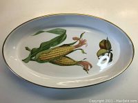 Oval white casserole dish with corn and fruit hand-painted design, gold trim around edge, on a beige surface.