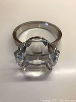 Front view of a large ring with a clear multi-faceted glass stone set in claw prongs on a silver-tone band.