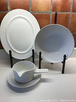 Photo showing all three items in the set: oval platter, serving bowl, gravy boat with saucer, all white with ridged detailing on the rims, displayed on black stands.