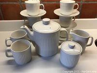 Front view of the set showing stacked six espresso/tea cups on saucers, two mugs, lidded teapot, creamer, and lidded sugar bowl all white ceramic with vertical ridged pattern.