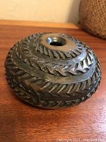 Dark textured ceramic vessel showing rugged geometric carvings, circular top opening, rough finish with chevron patterning.