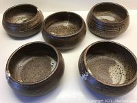 Five rustic clay bowls with sandy speckling, rich chocolate and cinnamon tones with cream drips, arranged on a white surface showing their exterior ridged texture.