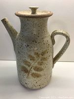 Front view of the studio pottery pot showing the body, handle, spout, and lid with speckled cream and brown glaze and engraved leaf motif.