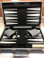 Open backgammon game board showing grey and black playing surface, cream and black points, two grey dice cups, two pairs of dice (black and cream), doubling cube, and black and cream playing pieces organized on the board.