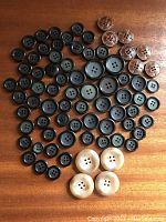 Top view showing black buttons arrayed from small to large sizes, 6 brown leather buttons arranged in a group, 4 large cream-colored buttons, and tortoise shell colored buttons.