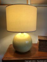 Full view of the lamp showing round yellow ceramic base glowing warmly under an off-white cylindrical shade with blue trim on top edge.