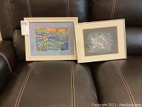 Two framed art pieces on a dark leather couch, one wooden framed colorful fishing scene and one metal framed abstract image.
