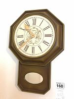 Front view of one octagonal plastic wall clock with white face, Roman numerals, and lower section with oval window