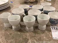 Set of 4 white egg cups and 7 white teacups with blue scenic prints displayed on countertop